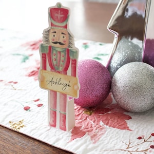 May include: A watercolor-style Christmas nutcracker place card holder with the name "Ashleigh" on a banner. The nutcracker is red, green, and white. A silver Christmas tree and two glittery ornaments, one pink and one silver, are in the background.