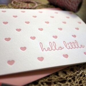 May include: A white card with a pink heart pattern and the text "hello little one" in pink lettering.