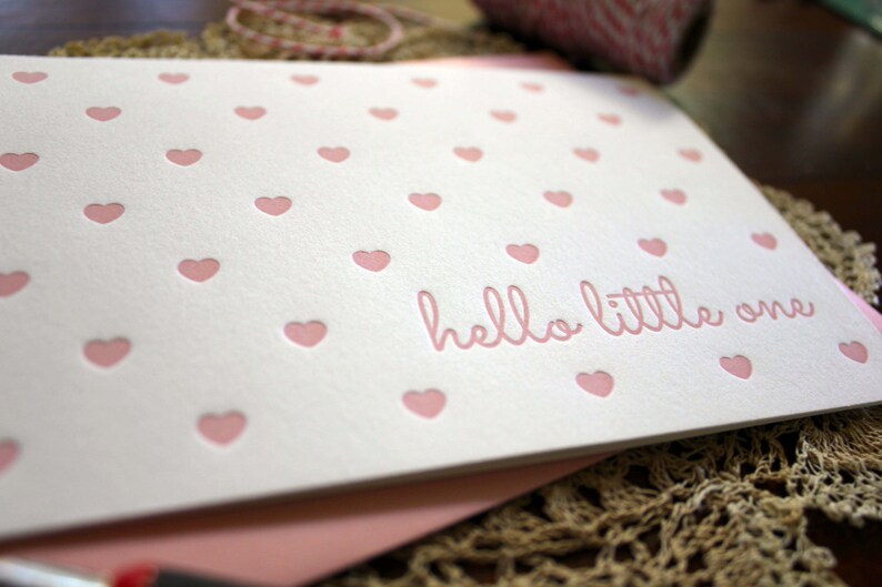 May include: A white card with a pink heart pattern and the text "hello little one" in pink lettering.