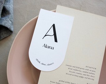 Könnte beinhalten: Eine weiße Tischkarte mit einem schwarzen Buchstaben "A" und dem Namen "Alana" darauf gedruckt. Der Text "drink, dine, dance" ist unten auf der Karte gedruckt.