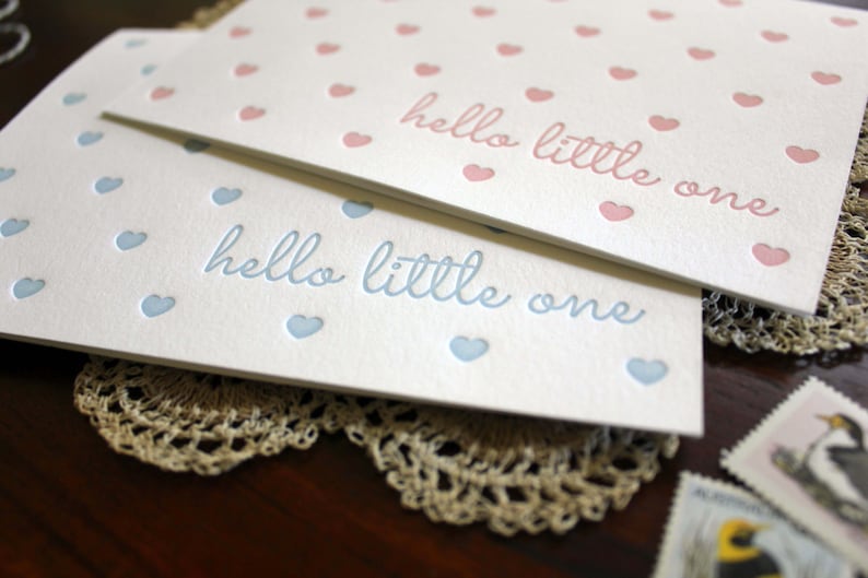 May include: Two white cards with the text "hello little one" printed in blue and pink. The cards are decorated with small blue and pink hearts.