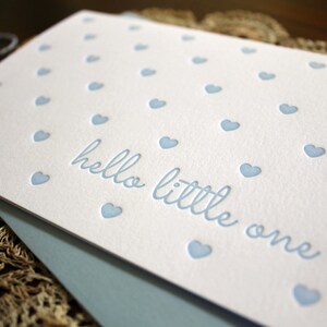 May include: A white card with a blue background and a pattern of small blue hearts. The card says "hello little one" in a blue script font.