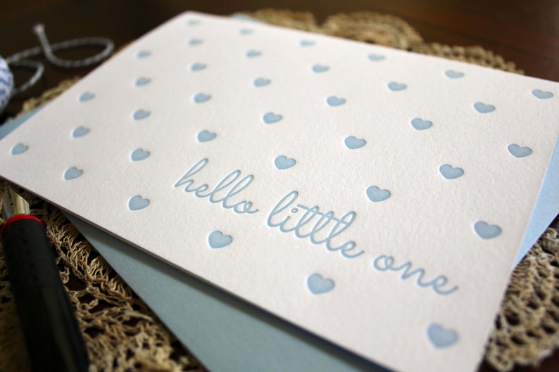 May include: A white card with a blue background and a pattern of small blue hearts. The card says "hello little one" in a blue script font.