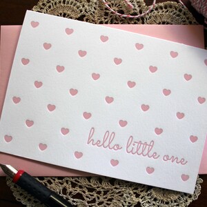 May include: A white card with a pink heart pattern and the text "hello little one" in pink. The card is on a white lace doily.