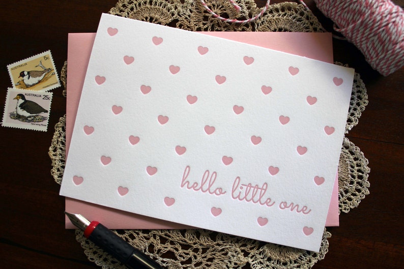 May include: A white card with a pink heart pattern and the text "hello little one" in pink. The card is on a white lace doily.