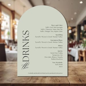 May include: A menu card with an arched top, listing drinks such as beer, cider, sparkling wine, spirits, and non-alcoholic beverages. The menu is on a wooden table in a restaurant setting. The text "DRINKS" is vertically aligned on the left side.