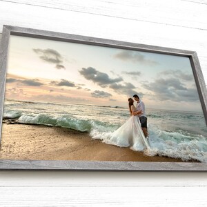 Pode incluir: Um casal em trajes de casamento est&aacute; em uma praia com uma onda quebrando atr&aacute;s deles. O casal est&aacute; se abra&ccedil;ando e olhando um para o outro. O c&eacute;u &eacute; de um tom rosa e laranja suave, e o oceano &eacute; azul profundo.