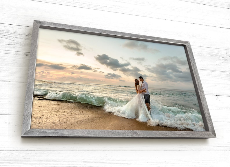 Pode incluir: Um casal em trajes de casamento est&aacute; em uma praia com uma onda quebrando atr&aacute;s deles. O casal est&aacute; se abra&ccedil;ando e olhando um para o outro. O c&eacute;u &eacute; de um tom rosa e laranja suave, e o oceano &eacute; azul profundo.