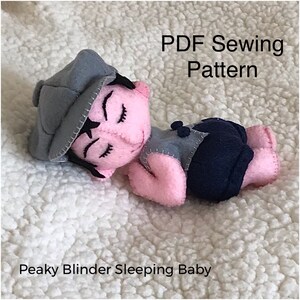 Può includere: Bambola in feltro fatta a mano a forma di bambino che dorme, con berretto grigio e pantaloni blu scuro. La bambola ha la pelle rosa e gli occhi chiusi. Il testo "PDF Sewing Pattern" e "Peaky Blinder Sleeping Baby" sono visibili.