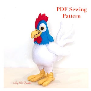 Op de afbeelding: Een handgemaakte vilten haan met een rode kam, blauwe kop en gele snavel en poten. Het lichaam is wit en de staartveren zijn ook wit. De afbeelding bevat de tekst "PDF Sewing Pattern" en "My 3D Rooster".