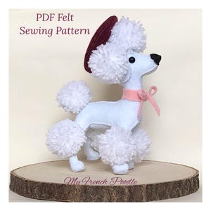 May include: A white felt French poodle with a burgundy beret and pink bow. The poodle has white pom-poms for fur and is standing on a wooden base. The text "My French Poodle" is visible on the base.