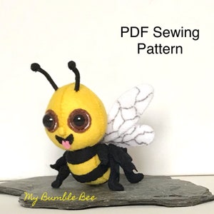 May include: A PDF sewing pattern for a felt bumble bee toy. The bee is yellow with black stripes, large brown eyes, and a pink tongue sticking out. The bee has white wings with black veins.