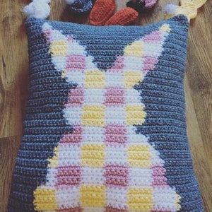 May include: A blue crocheted pillow with a pink and yellow checkered bunny silhouette. The pillow is decorated with small crocheted bunnies and carrots.