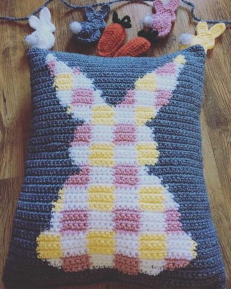 May include: A blue crocheted pillow with a pink and yellow checkered bunny silhouette. The pillow is decorated with small crocheted bunnies and carrots.