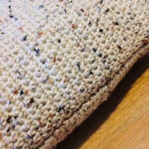 May include: Close-up of a cream-colored crocheted pillow with flecks of brown, black, and blue throughout the fabric.