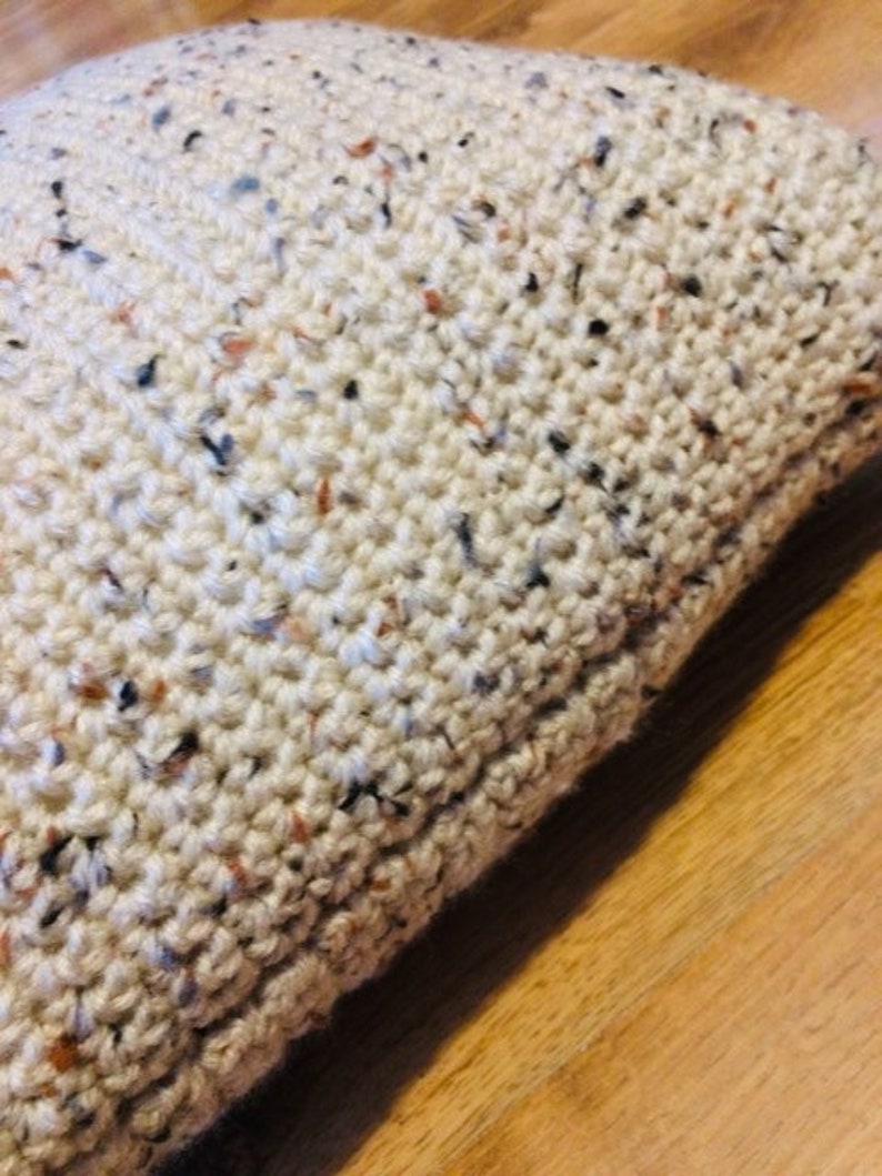 May include: Close-up of a cream-colored crocheted pillow with flecks of brown, black, and blue throughout the fabric.