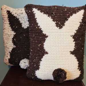 May include: Two crocheted pillows in the shape of a bunny rabbit. One pillow is brown with white accents, and the other is white with brown accents. Both pillows have a brown pom-pom tail.