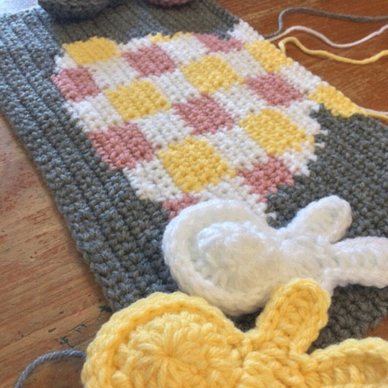May include: A crocheted blanket with a checkered pattern in pink, yellow, and white. The blanket is surrounded by crocheted flowers and a bunny in white.