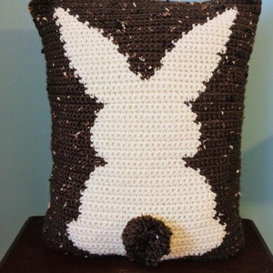 May include: A brown and white crocheted pillow shaped like a bunny rabbit. The bunny has a brown pom-pom tail.