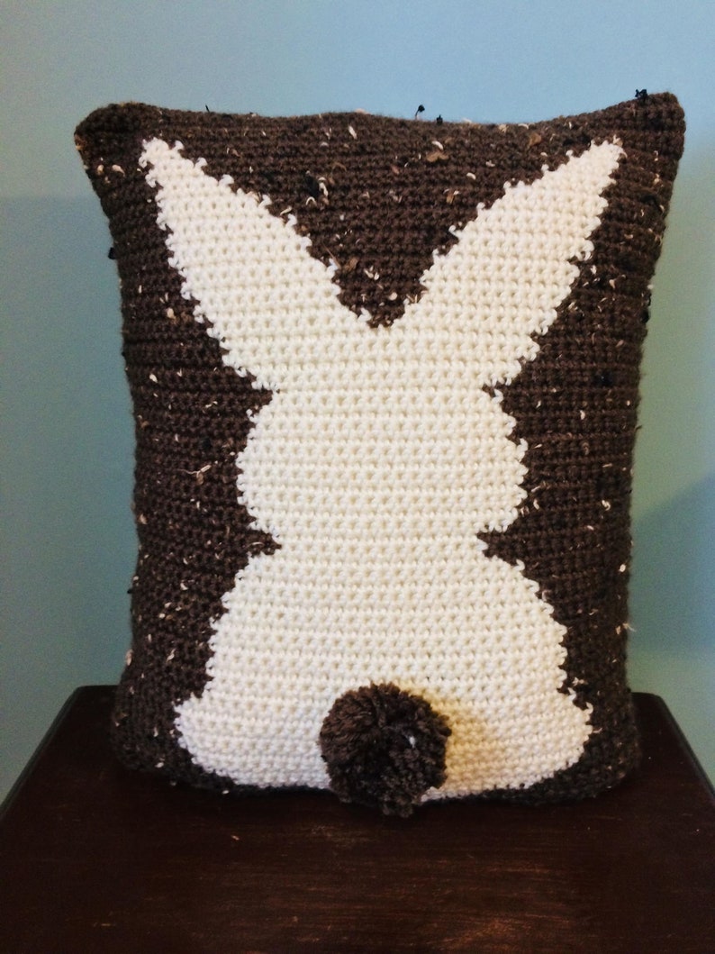 May include: A brown and white crocheted pillow shaped like a bunny rabbit. The bunny has a brown pom-pom tail.