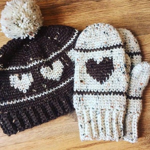 Op de afbeelding: Een gebreide muts van bruin en wit met een witte pompon en bijpassende gebreide wanten van bruin en wit. De muts en wanten hebben een hartvormig patroon.