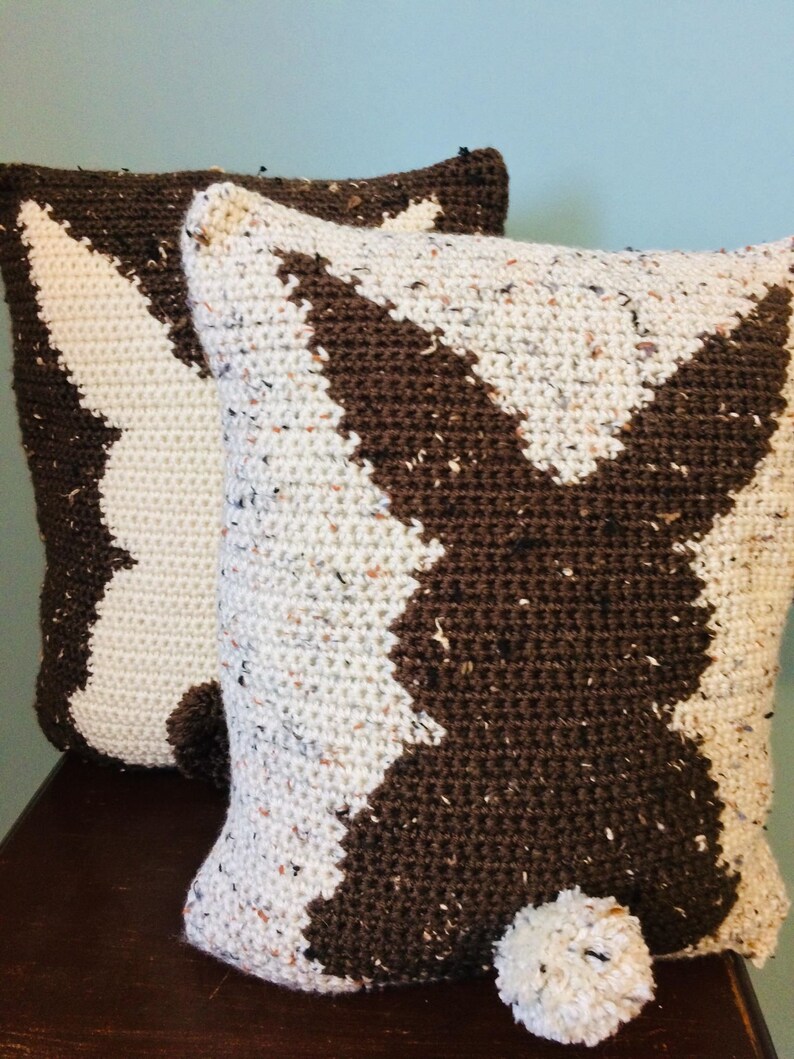 May include: Two crocheted pillows with a brown and white chevron pattern. One pillow has a brown pom-pom on the bottom.