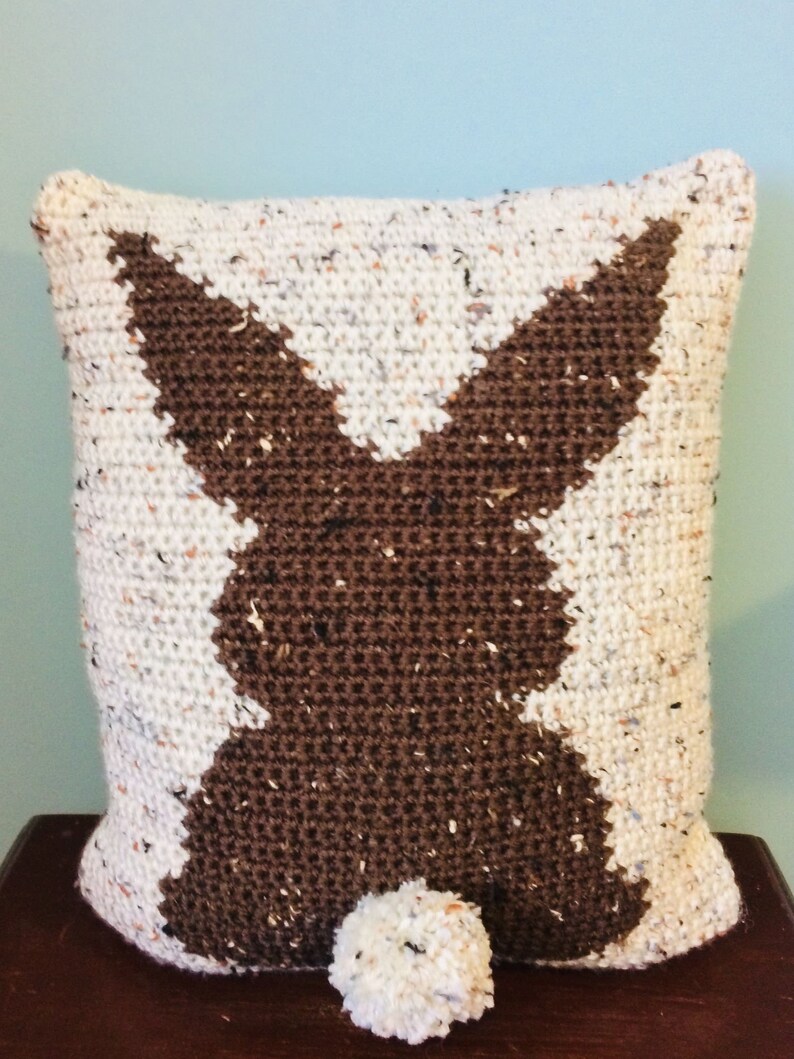 May include: A white and brown crocheted pillow with a bunny silhouette and a white pom-pom tail.
