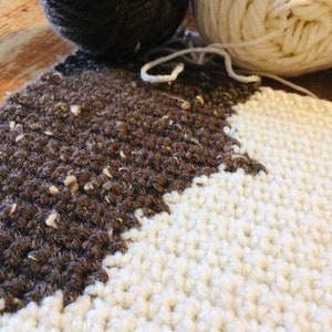 May include: Close-up of a brown and white crocheted blanket. The blanket is made with two different types of yarn, one brown and one white. The brown yarn has a speckled texture.