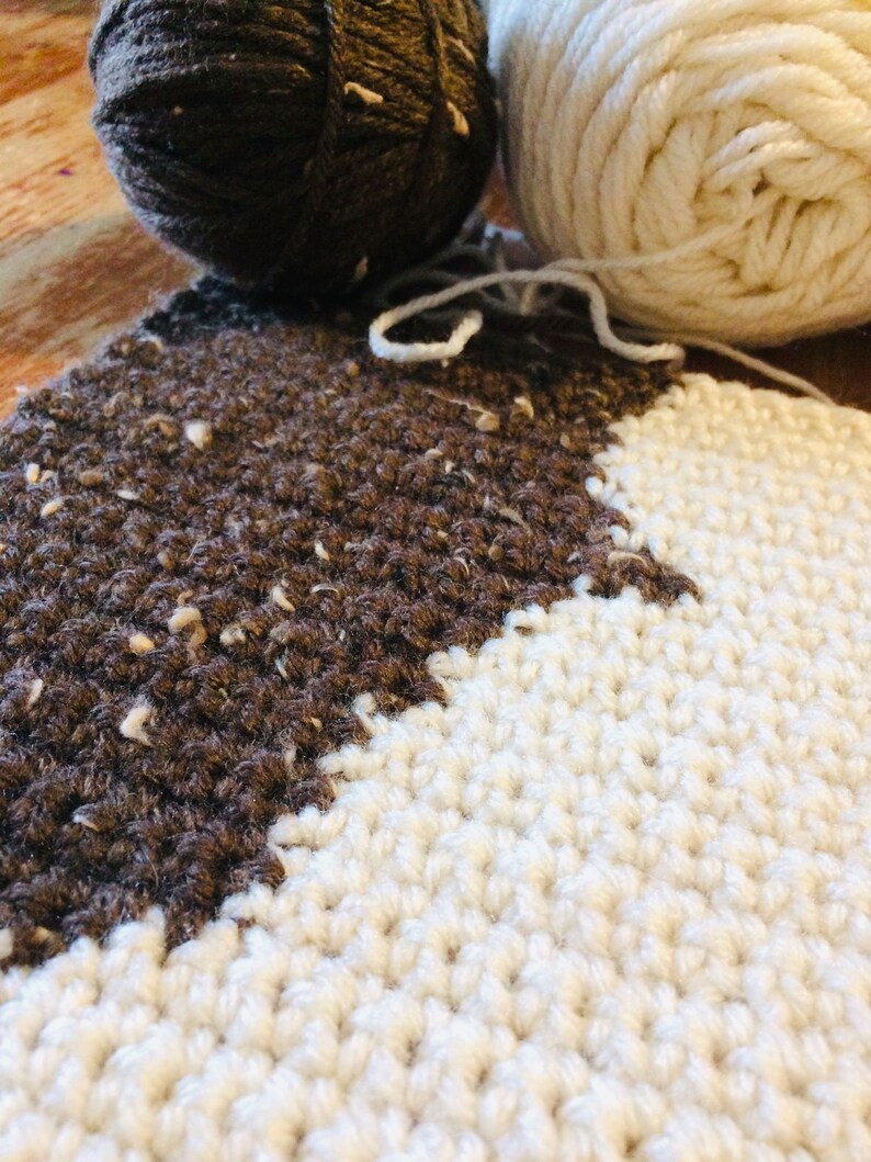 May include: Close-up of a brown and white crocheted blanket. The blanket is made with two different types of yarn, one brown and one white. The brown yarn has a speckled texture.