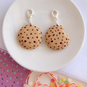 Puede incluir: Dos colgantes con forma de galleta con cierres plateados. Los colgantes son de color marrón claro con manchas marrones oscuras y caras sonrientes. Un colgante tiene un mordisco. Los colgantes están sobre un plato blanco.