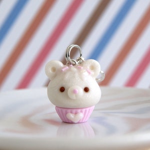 Könnte beinhalten: Ein weißer Cupcake-Anhänger mit einem rosa Herz und einem Bärenkopf. Der Cupcake befindet sich auf einem weißen Teller mit gestreiftem Hintergrund.