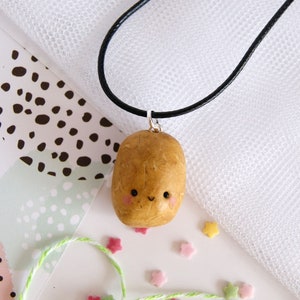 May include: A brown potato shaped pendant with a happy face on a black cord necklace. The pendant is made of polymer clay and has a glossy finish.
