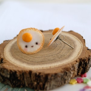 May include: A pair of cute, handmade earrings shaped like fried eggs with smiling faces. The earrings are made of polymer clay and have a light brown colour with a yellow yolk in the centre. The earrings are on a wooden surface.
