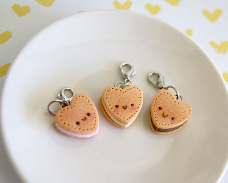 May include: Three heart-shaped charm pendants resembling sandwich cookies with smiling faces. The cookies are tan with pink, white, and brown fillings. Each charm has a silver-colored clasp.