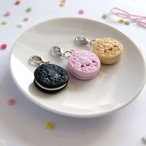 Op de afbeelding: Drie miniatuurkoekjescharms met lachende gezichten. De koekjes zijn zwart, roze en beige met witte vulling. De charms zijn bevestigd aan zilveren springringen.