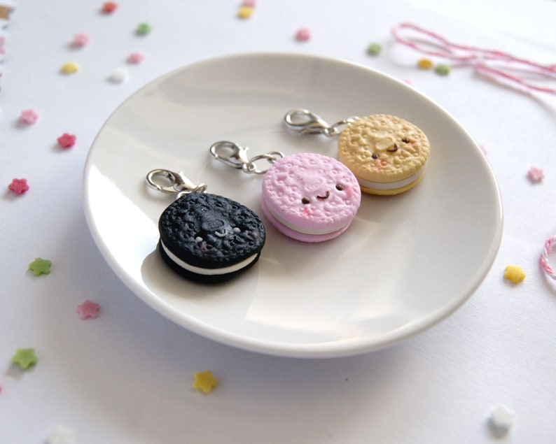 Op de afbeelding: Drie miniatuurkoekjescharms met lachende gezichten. De koekjes zijn zwart, roze en beige met witte vulling. De charms zijn bevestigd aan zilveren springringen.