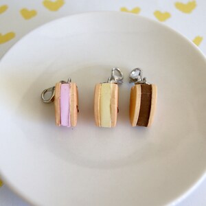 May include: Three miniature ice cream sandwich charms with silver-colored findings. Each charm features a different colored filling: pink, yellow, and brown. The charms are displayed on a white plate against a background with yellow heart shapes.