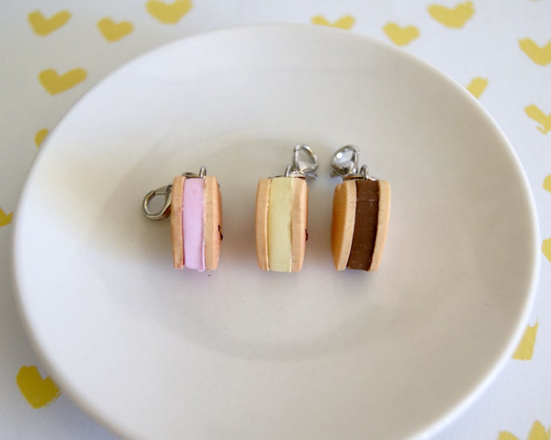 May include: Three miniature ice cream sandwich charms with silver-colored findings. Each charm features a different colored filling: pink, yellow, and brown. The charms are displayed on a white plate against a background with yellow heart shapes.