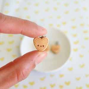 May include: A small, heart-shaped charm with a smiling face and dotted border. The charm is held between fingers, with a second charm visible in the background on a white plate. The background has a pattern of yellow hearts.