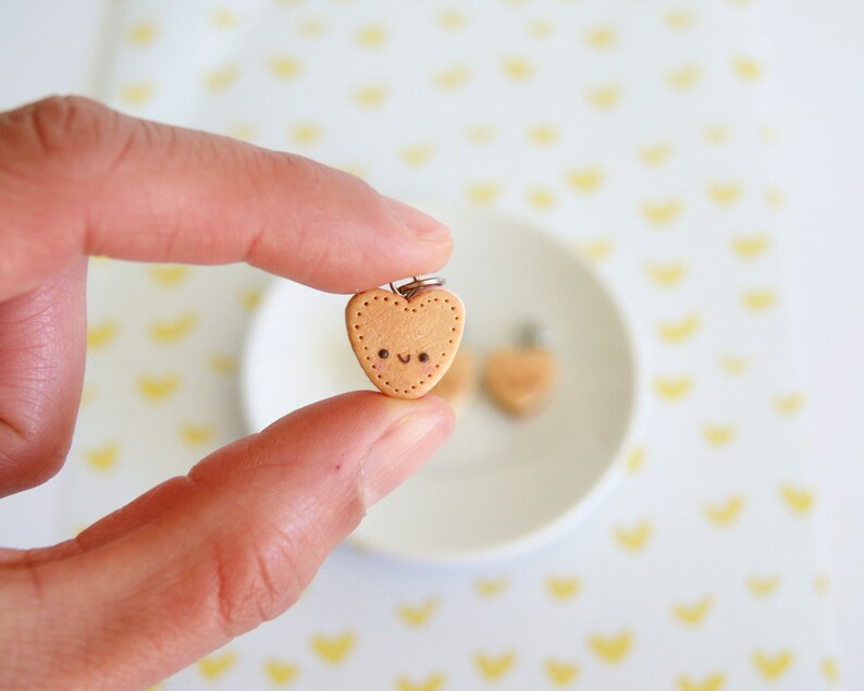 May include: A small, heart-shaped charm with a smiling face and dotted border. The charm is held between fingers, with a second charm visible in the background on a white plate. The background has a pattern of yellow hearts.