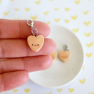 May include: A light peach-colored heart-shaped charm with a smiling face and dotted border. The charm is attached to a silver-colored clasp. A second charm is visible on a white plate.