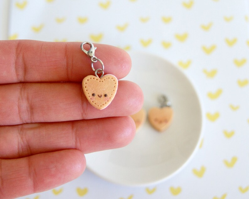 May include: A light peach-colored heart-shaped charm with a smiling face and dotted border. The charm is attached to a silver-colored clasp. A second charm is visible on a white plate.