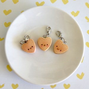 May include: Three heart-shaped charms with smiling faces and silver clasps rest on a white plate. The charms are light brown with pink accents and small black dots for eyes and a stitched border. The background is white with yellow heart-shaped brushstrokes.