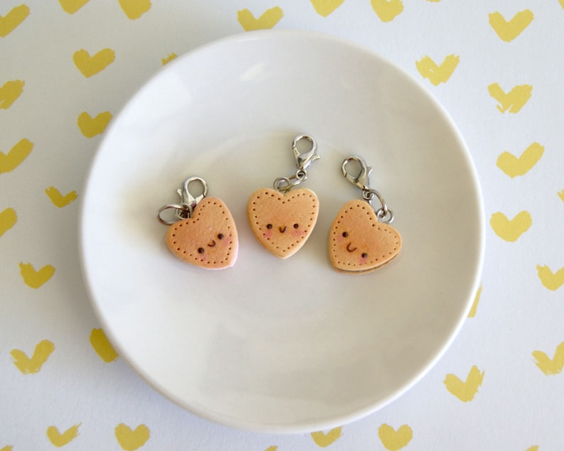 May include: Three heart-shaped charms with smiling faces and silver clasps rest on a white plate. The charms are light brown with pink accents and small black dots for eyes and a stitched border. The background is white with yellow heart-shaped brushstrokes.