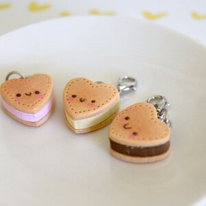 May include: Three heart-shaped charms resembling ice cream sandwiches. Each charm has a light brown exterior with a dotted border and a smiling face. The fillings are pink, yellow, and brown, and each charm has a silver clasp.