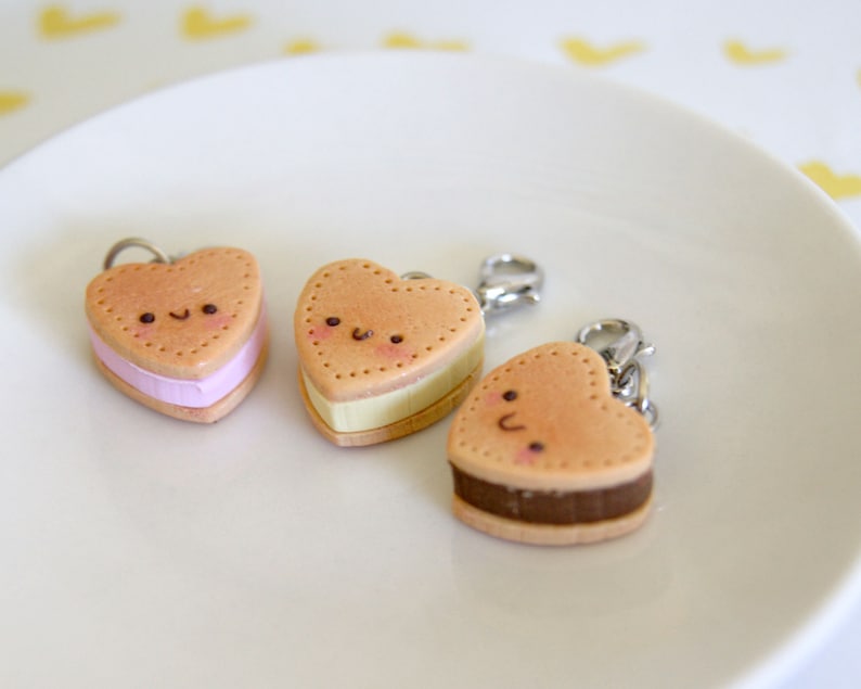 May include: Three heart-shaped charms resembling ice cream sandwiches. Each charm has a light brown exterior with a dotted border and a smiling face. The fillings are pink, yellow, and brown, and each charm has a silver clasp.