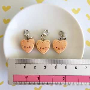 May include: Three heart-shaped charms with smiling faces and silver clasps. The charms are a light peach color with stitched edges. A ruler shows the charms are approximately 1 inch in width. The charms are on a white plate.