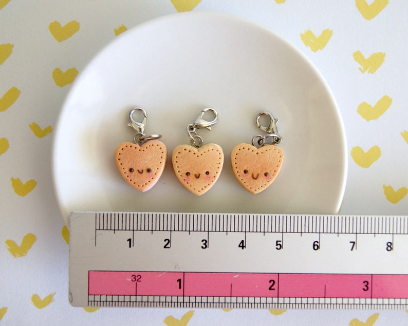 May include: Three heart-shaped charms with smiling faces and silver clasps. The charms are a light peach color with stitched edges. A ruler shows the charms are approximately 1 inch in width. The charms are on a white plate.
