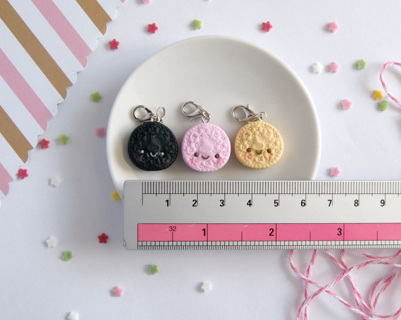 Op de afbeelding: Drie kleine bedels in de vorm van koekjes met vrolijke gezichten. De koekjes zijn zwart, roze en geel. De bedels liggen op een wit bord met een liniaal voor de schaal.