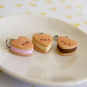 May include: Three heart-shaped keychains resembling ice cream sandwiches. Each keychain has a light brown cookie exterior with a different colored filling: pink, yellow, and brown. The keychains have smiling faces and are on a white plate.